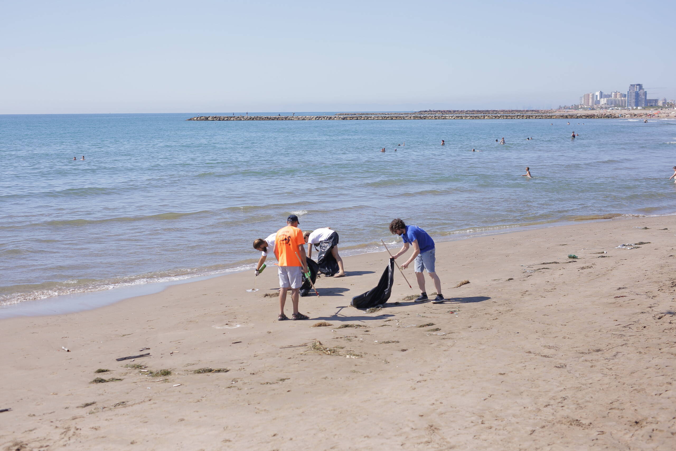 La imagen muestra una escena en una playa durante una actividad de limpieza ambiental. En primer plano, hay cuatro personas agachadas recogiendo residuos en la arena, utilizando bolsas grandes de basura. Visten ropa informal; una persona lleva una camiseta naranja, otra blanca, una azul y otra más al fondo con camiseta clara. El mar está en calma y ocupa el fondo de la imagen, con algunas personas bañándose o caminando en la orilla. El cielo está despejado y, a lo lejos, se distinguen edificios costeros y un espigón. La imagen transmite una acción colectiva de cuidado del medio ambiente en un entorno marino.