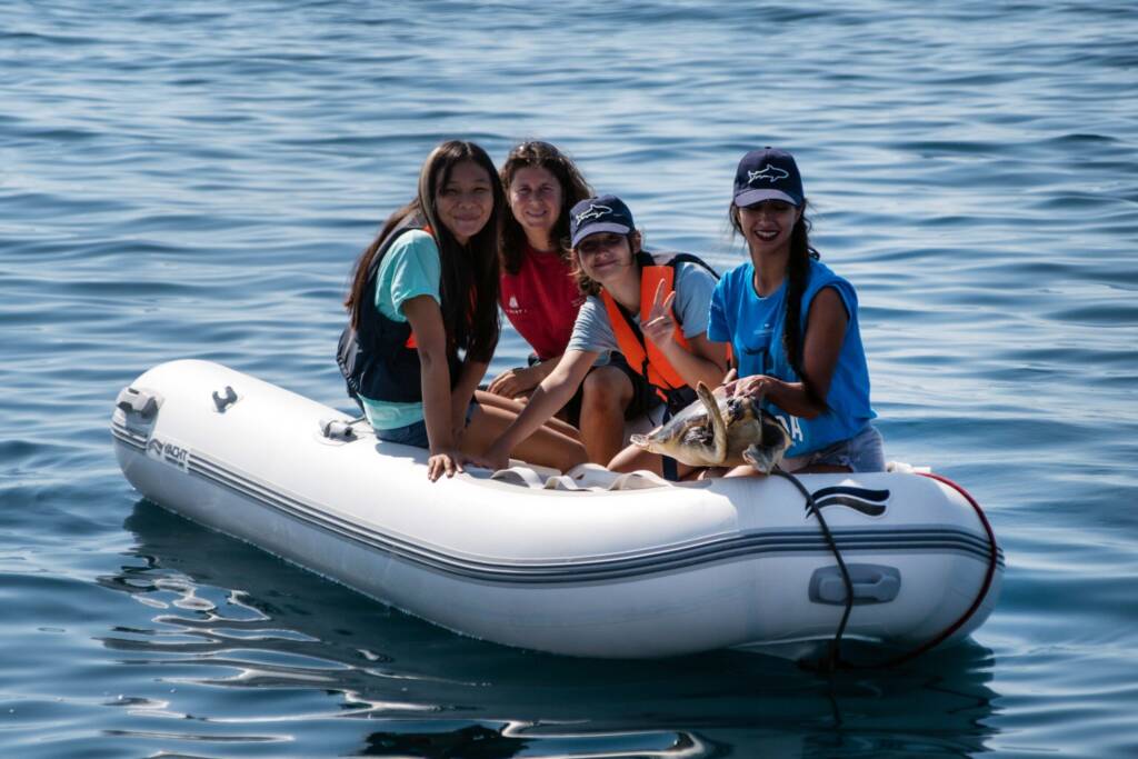 Cuatro jóvenes y una educadora, con chalecos salvavidas, están sentadas en una pequeña embarcación hinchable en el mar. Sostienen una tortuga marina, que va a ser devuelta al agua como parte de una actividad de conservación.