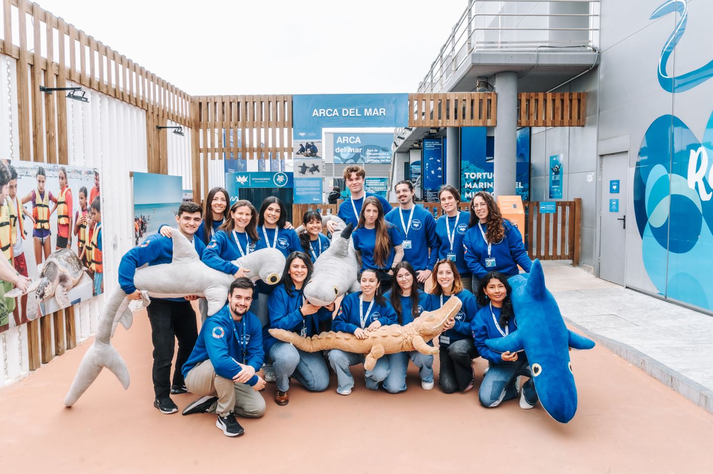 Grupo de jóvenes voluntarios posando sonrientes con peluches marinos frente al edificio del Arca del Mar del Oceanogràfic.