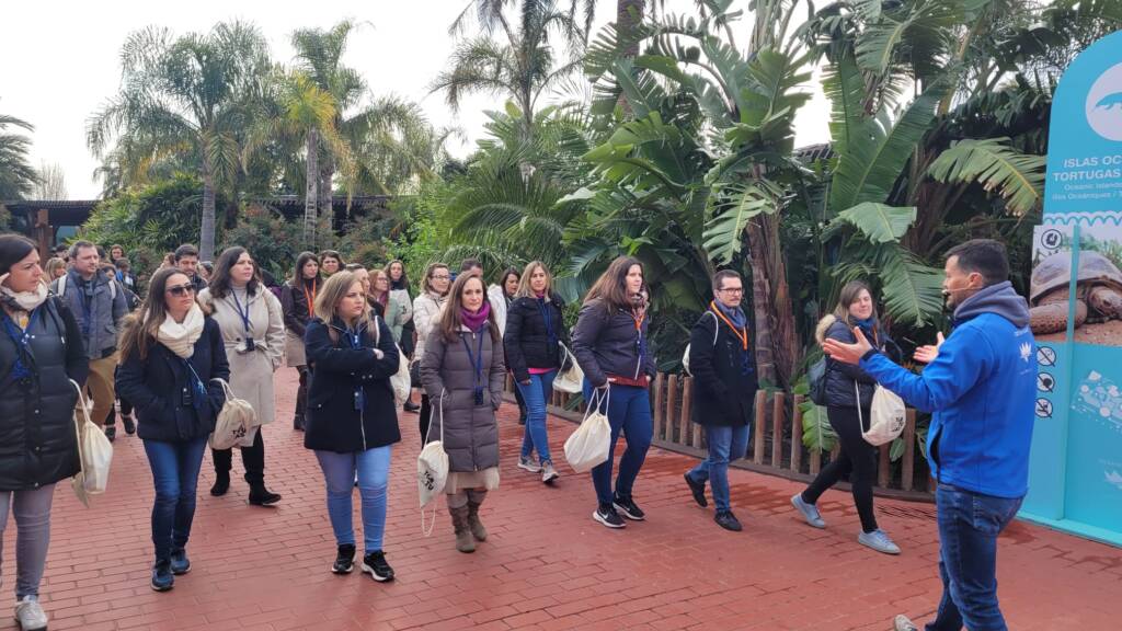 Un grupo de personas participa en una visita guiada al aire libre en el Oceanogràfic. Escuchan atentamente al guía, rodeados de vegetación tropical y junto a un cartel sobre tortugas marinas.