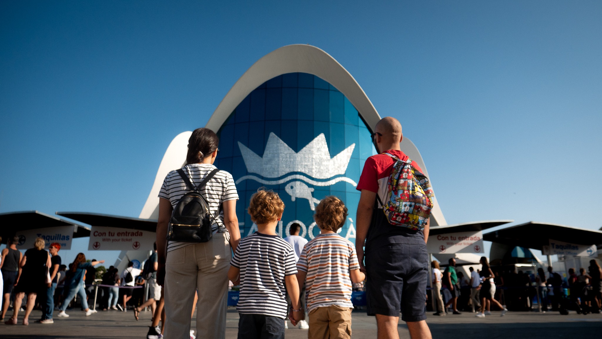 Una familia, compuesta por dos personas adultas y dos niños pequeños, se encuentra de espaldas frente a la entrada del Oceanogràfic de Valencia. Observan el edificio principal, de estructura curva y fachada azul con el logotipo del centro. A su alrededor, hay más visitantes haciendo fila para entrar, en un ambiente soleado y animado.