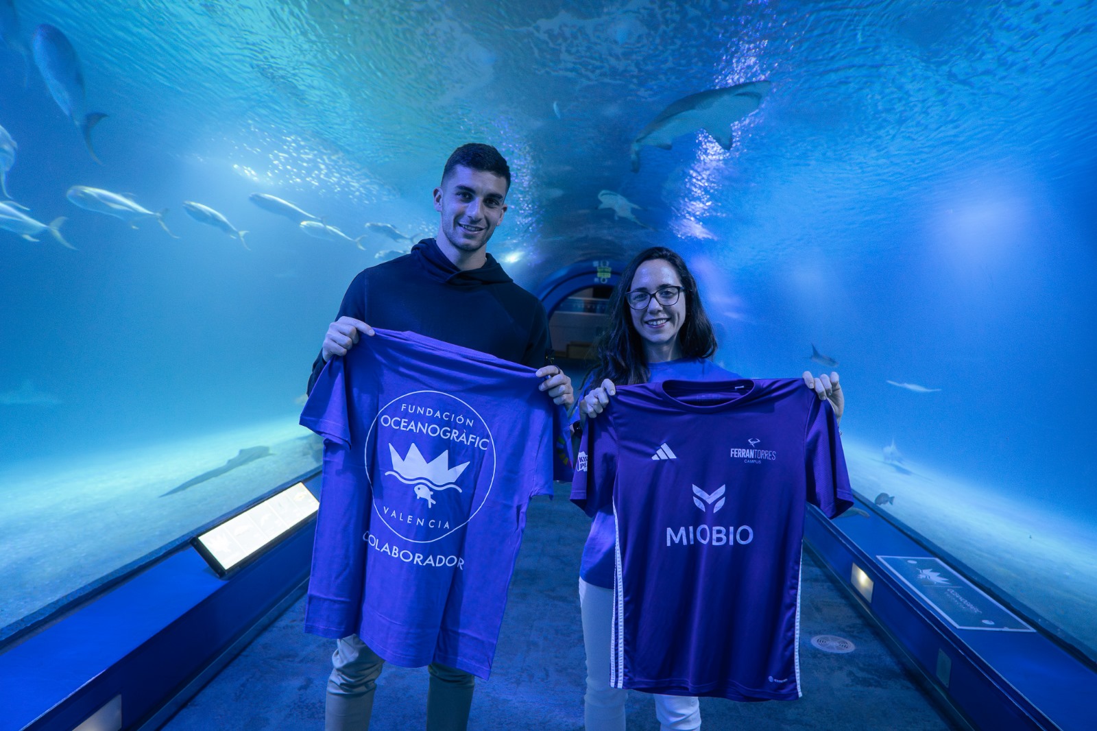 Ferran Torres y Alicia Borque, divulgadora de la Fundación Oceanogràfic, están de pie dentro de un túnel submarino del Oceanogràfic de Valencia, en la instalación Océanos del Oceanogràfic. Ambos sonríen y sostienen camisetas moradas: él muestra una con el logo de la Fundación Oceanogràfic, y ella otra del campus de fútbol de Ferran Torres, con el logotipo de la marca “MIOBIO”.